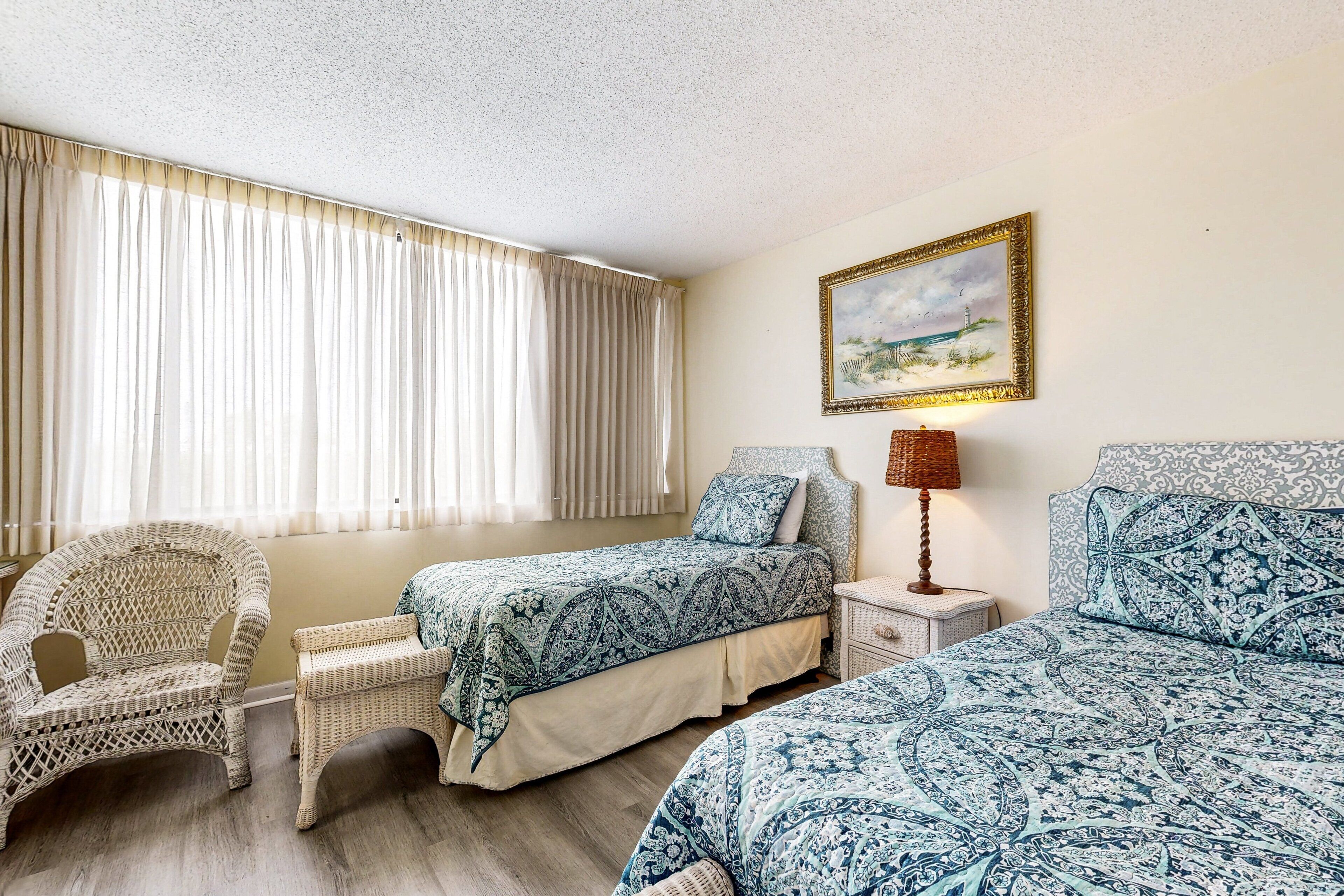 A room with two beds, a wicker chair, a nightstand with a lamp, and a painting on the wall.