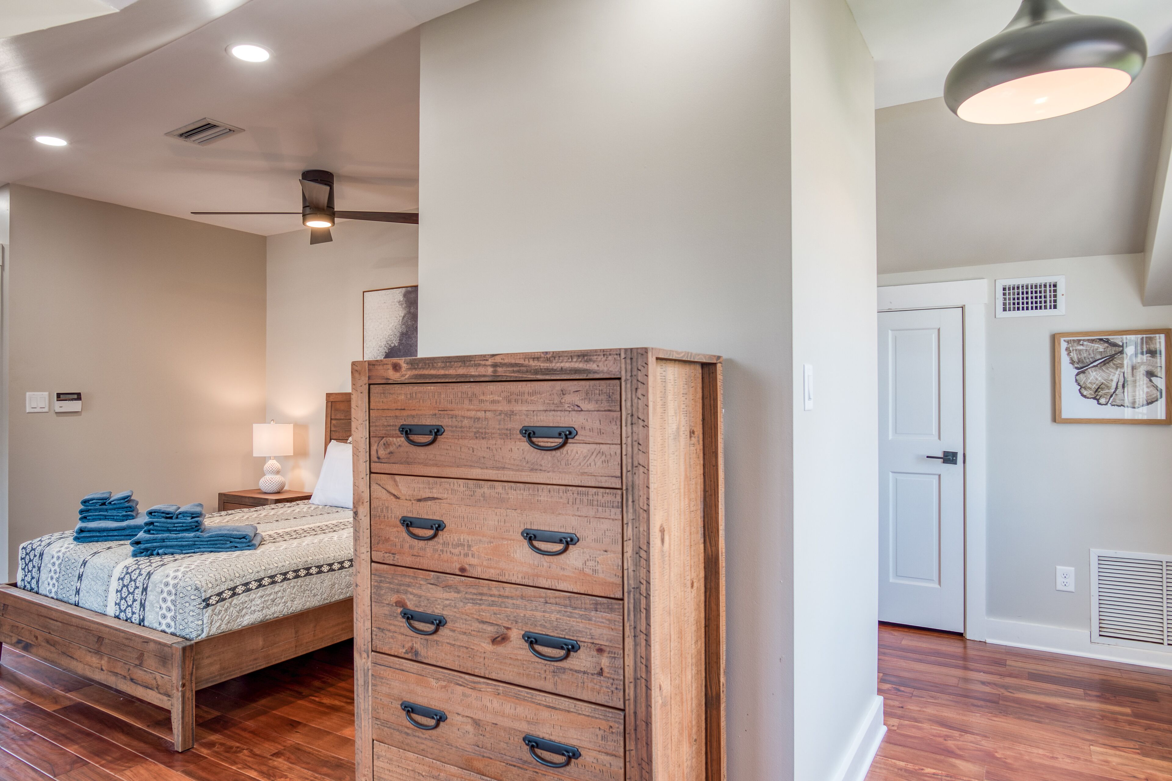A bedroom with a wooden dresser, a bed with blue bedding, a ceiling fan, and a white door.