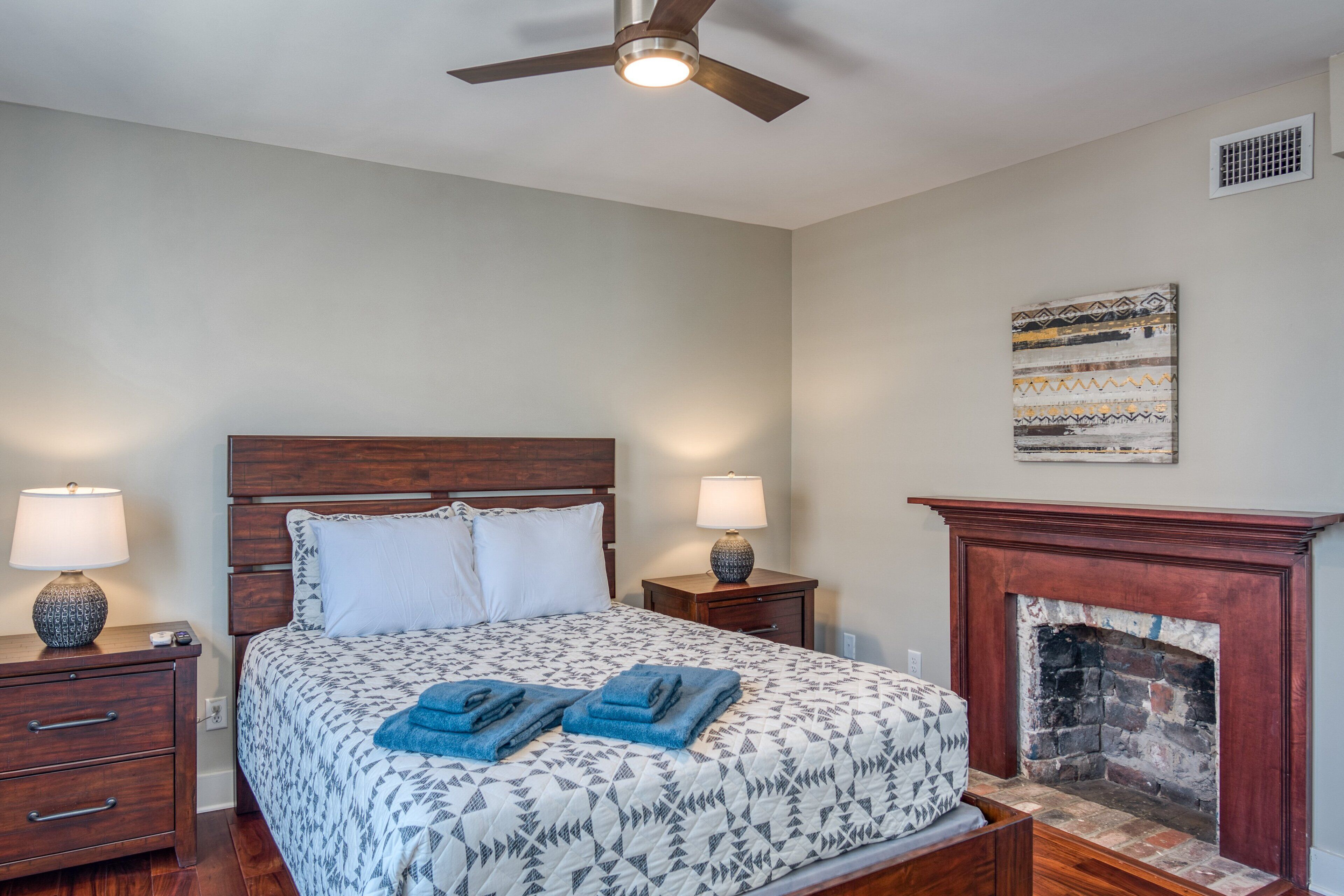 A bedroom with a bed, nightstands, a ceiling fan, a fireplace, and a wall-mounted artwork.