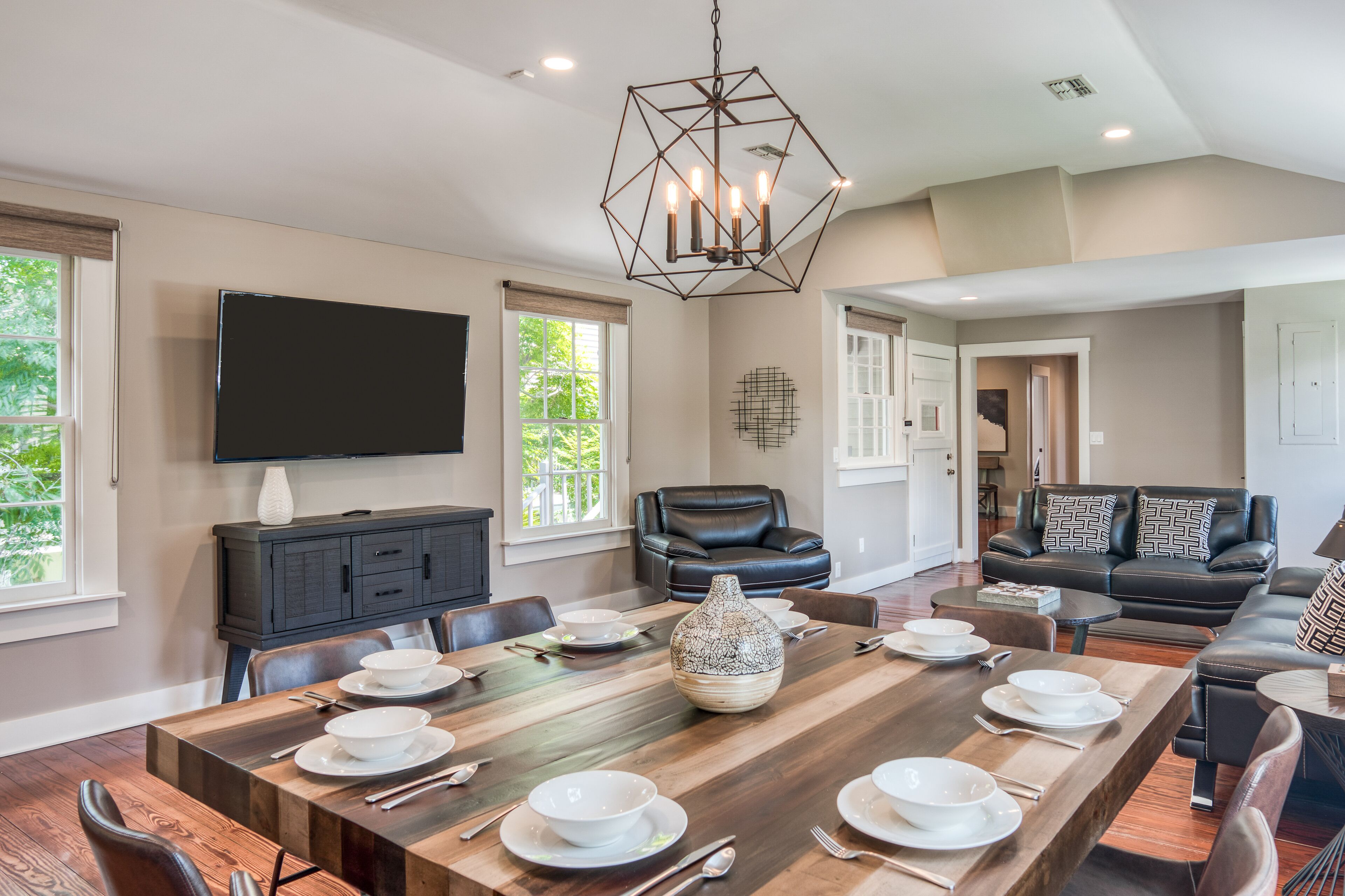 A spacious dining area with a long wooden table set for a meal, a black leather sofa, and a modern chandelier.