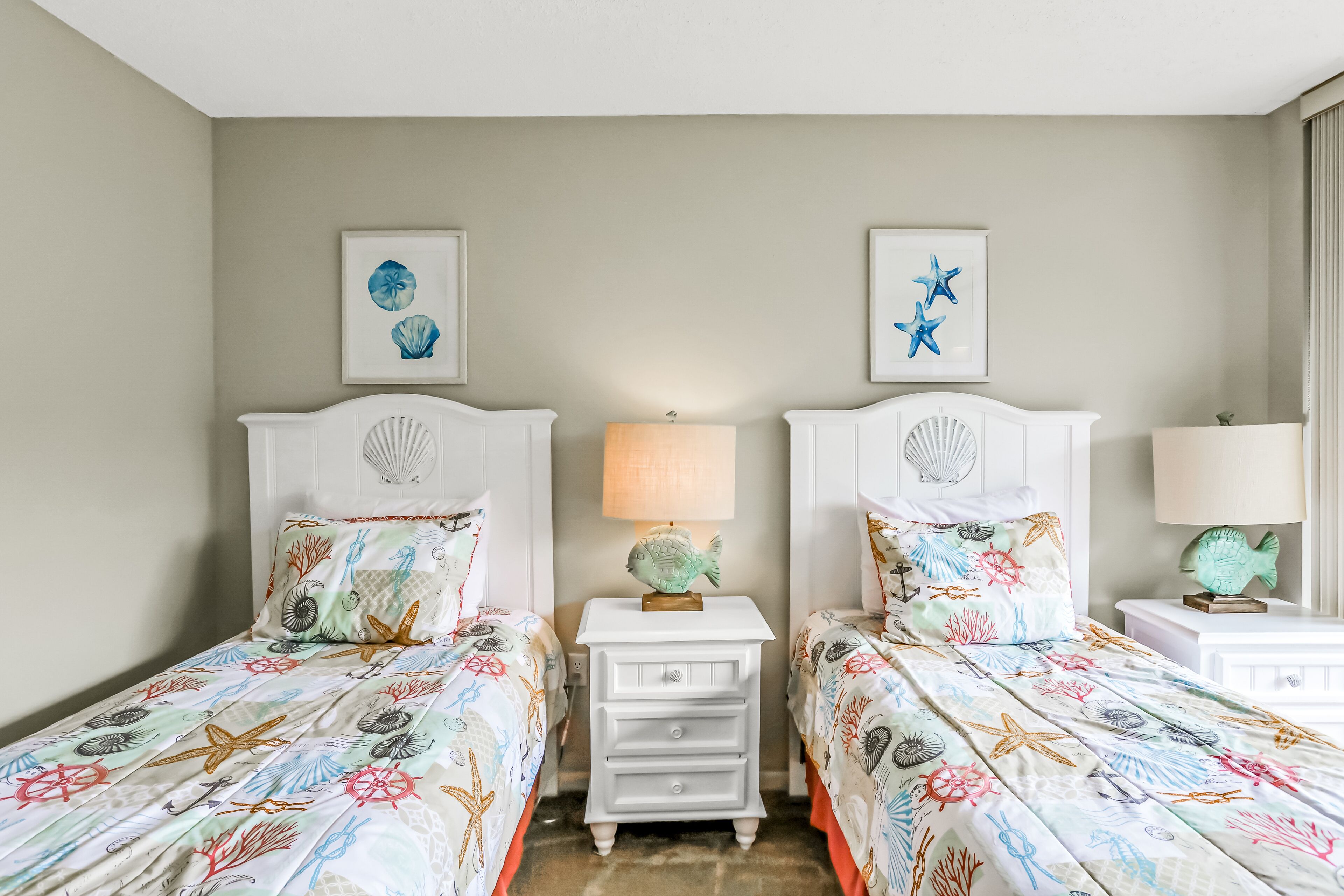A hotel room with two beds, each with a decorative headboard and matching pillows. There are wall-mounted lamps and framed artwork above the beds.