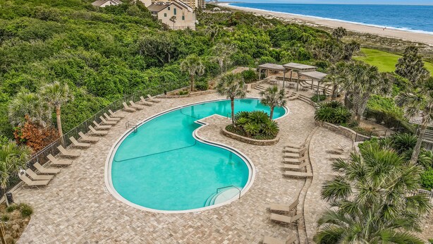 A resort with a large, curved swimming pool surrounded by lounge chairs and palm trees, overlooking a beach. - Charming Amelia Island Plantation Condo With Balcony, AC, Washer/dryer & Pool (Fernandina Beach)