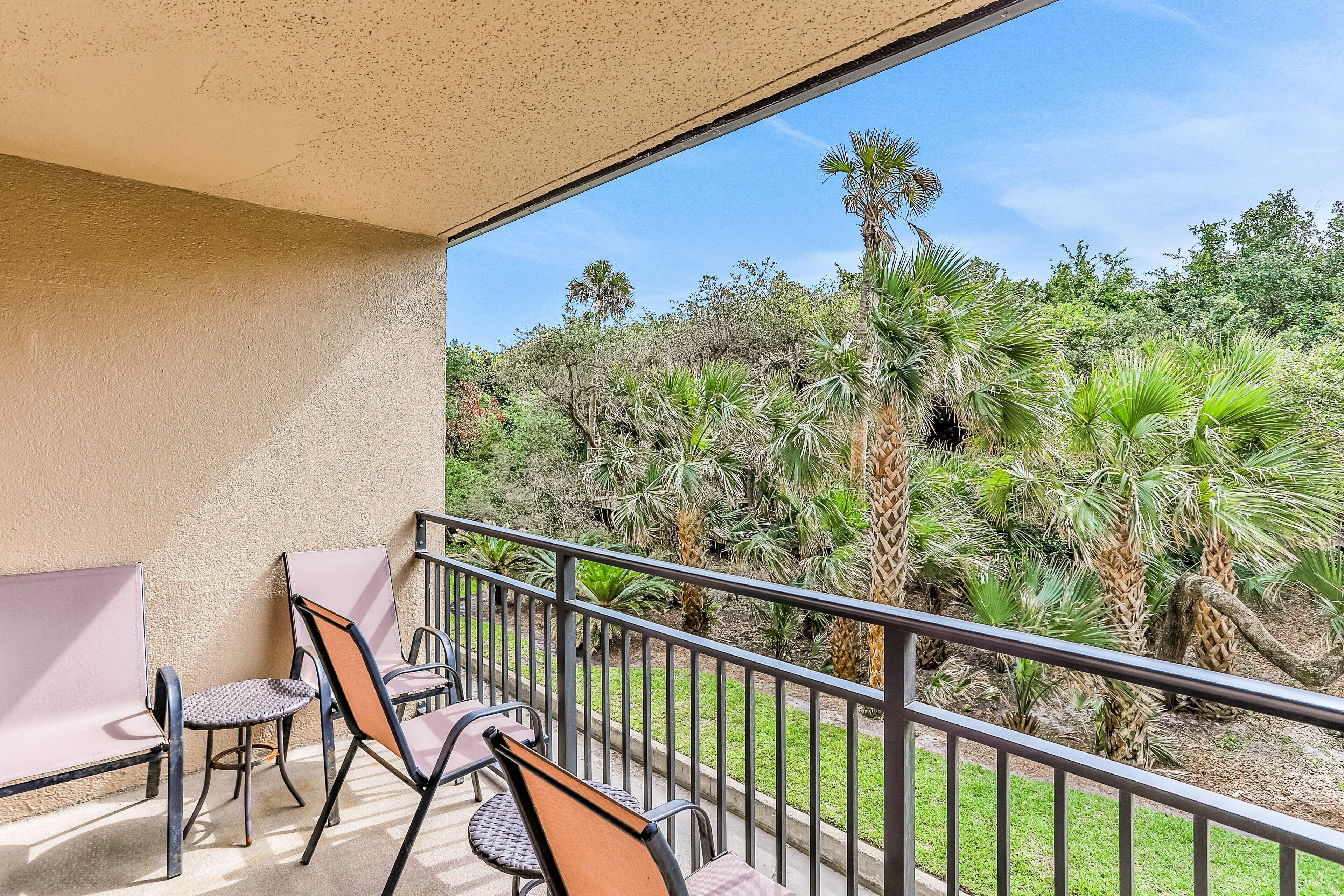 A balcony with two chairs and a table, overlooking a garden with various plants and trees.