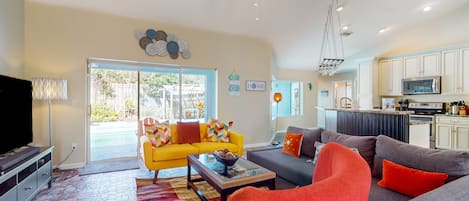 A modern living room with a yellow sofa, grey sofa, and red armchairs. There is a kitchen area with white cabinets and a microwave.