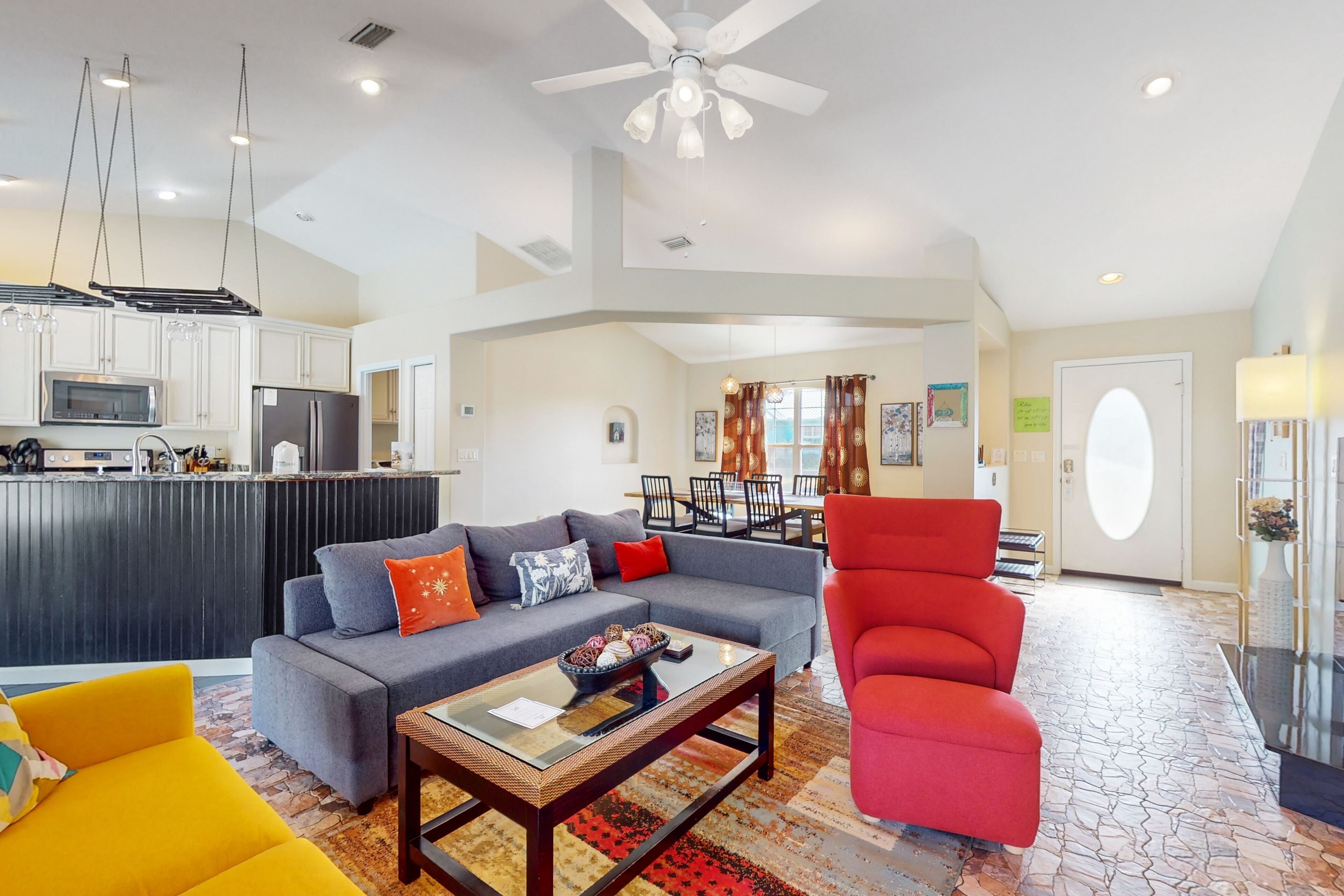 A spacious living area with a gray sofa, a red armchair, and a yellow armchair. There is a kitchen area with white cabinets and a microwave. A ceiling fan is visible, and the room has a tiled floor.