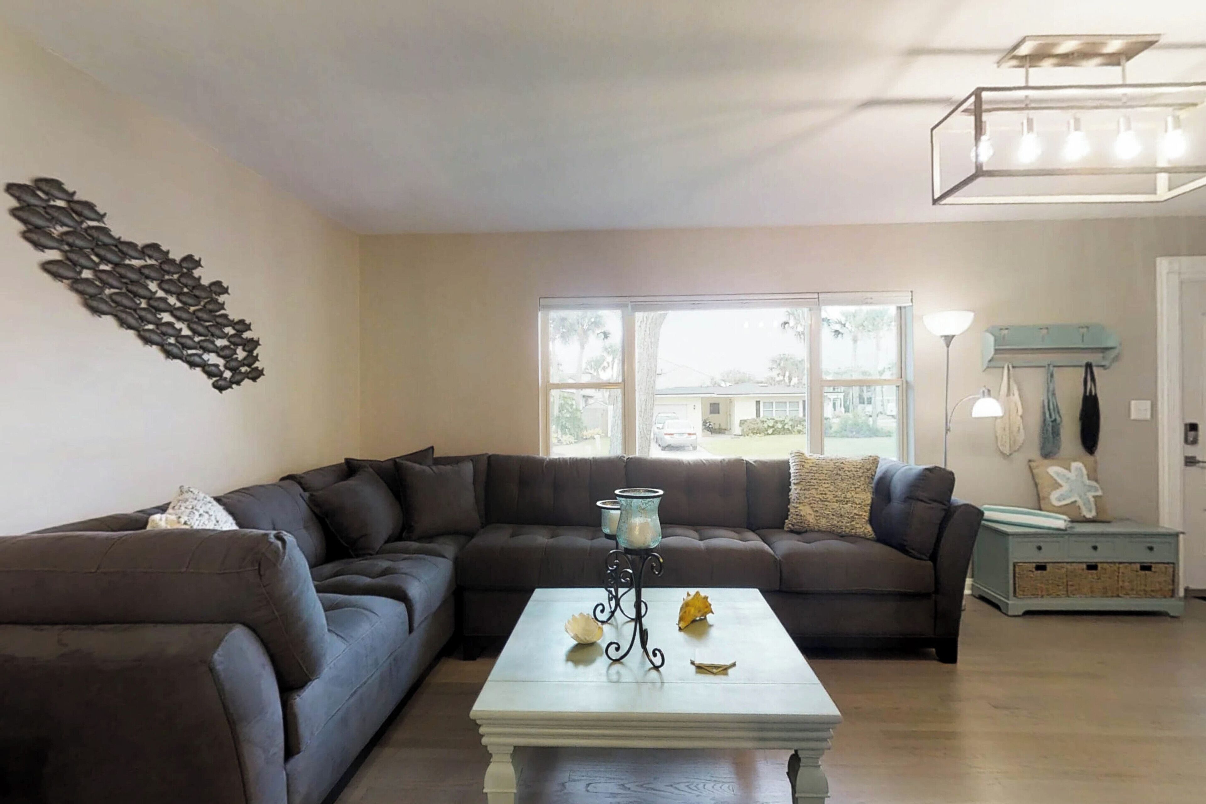 A living room with a sectional sofa, a white coffee table with decorative items, a wall art piece, and a window with a view of the outside.