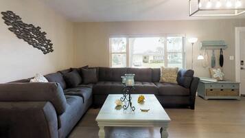 A living room with a sectional sofa, a white coffee table with decorative items, a wall art piece, and a window with a view of the outside.