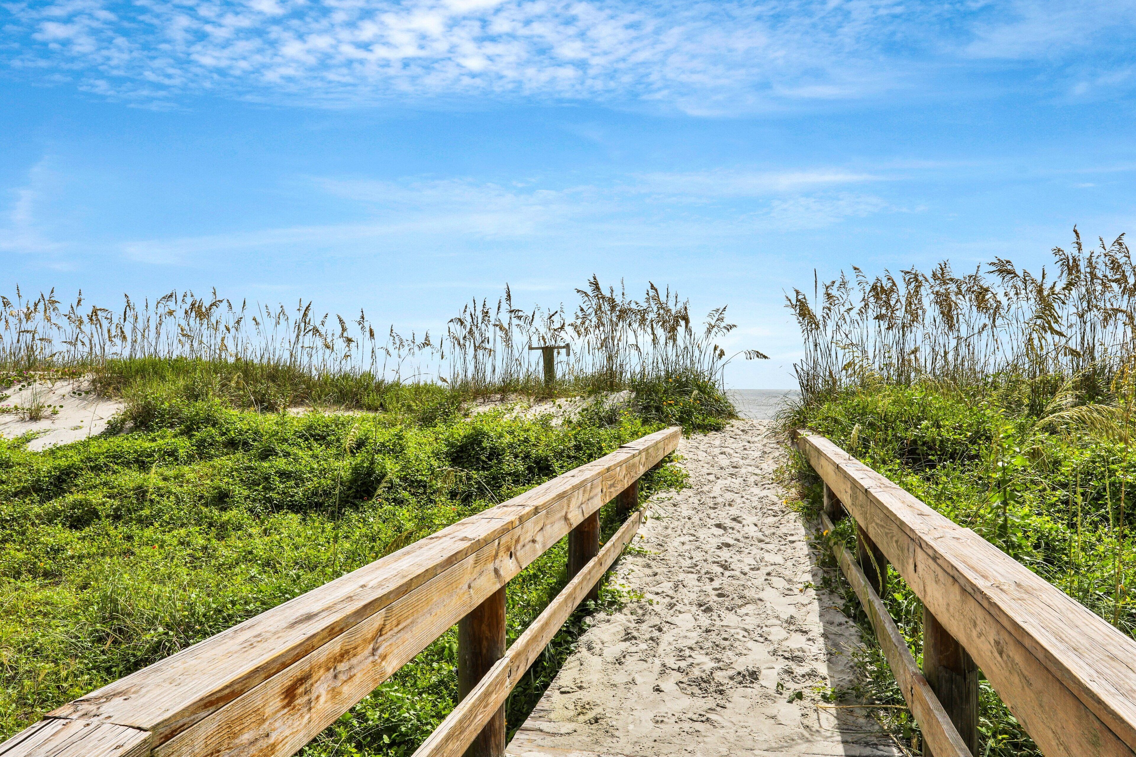 A wooden boardwalk leading to a beach with sand and grassy vegetation on either side.