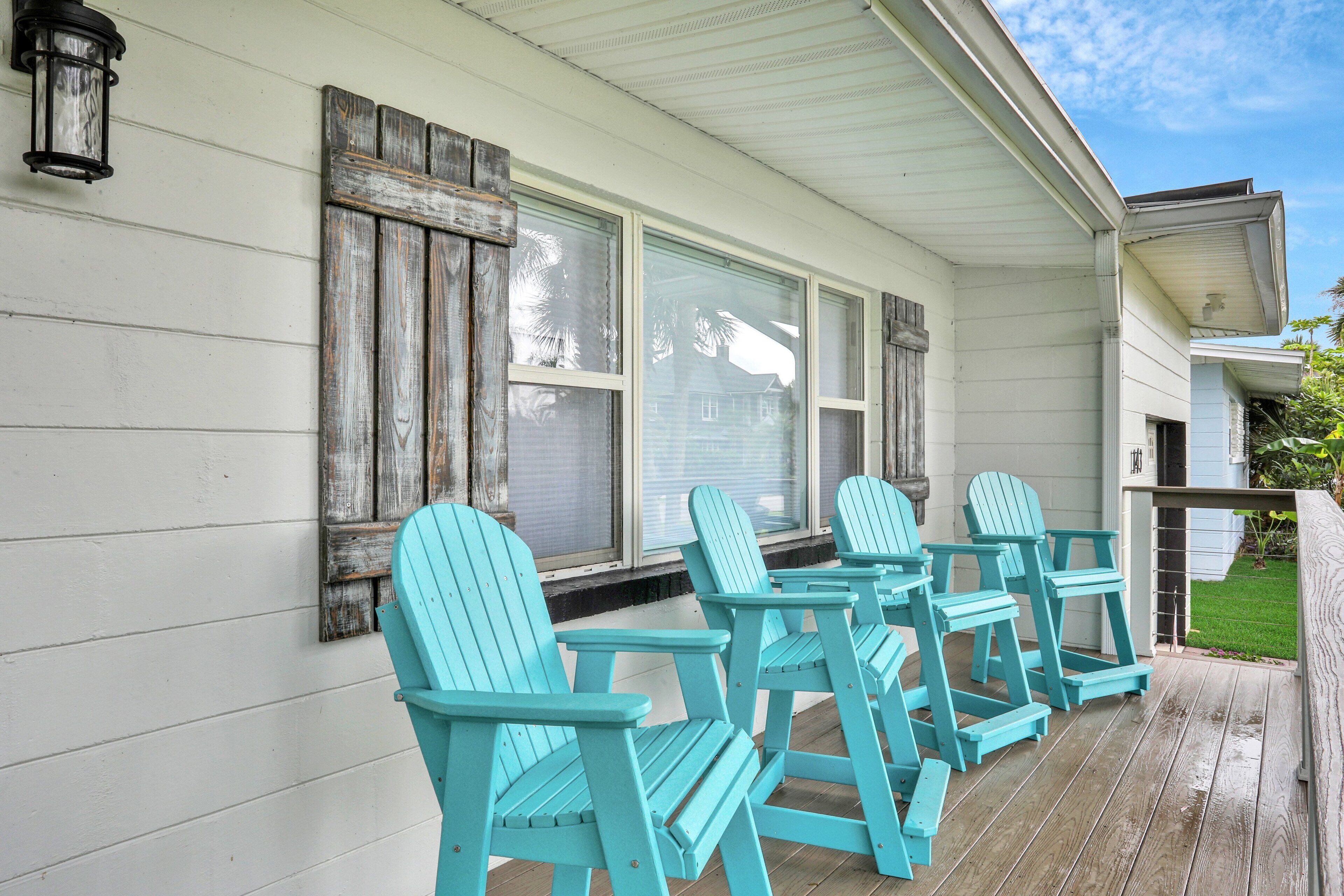 A porch with four turquoise Adirondack chairs, a white exterior wall, and a wooden deck.