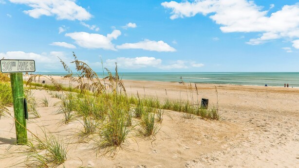 Een strand met een bord waarop '30th AVES' staat, en een zanderig landschap met schaarse begroeiing.