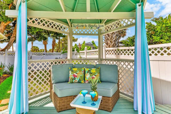 Ein überdachter Außenbereich mit einer Rattan-Couch, zwei bunten Kissen, einer Ablage mit zwei Getränken und einer Ananas, sowie einem Schild mit der Aufschrift 'Welcome to Beachy Jean'.