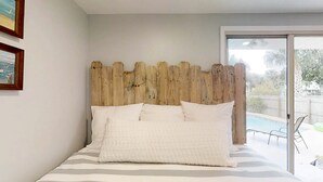 Een slaapkamer met een houten hoofdbord, een bed met gestreept beddengoed en uitzicht op een buitenzwembad.