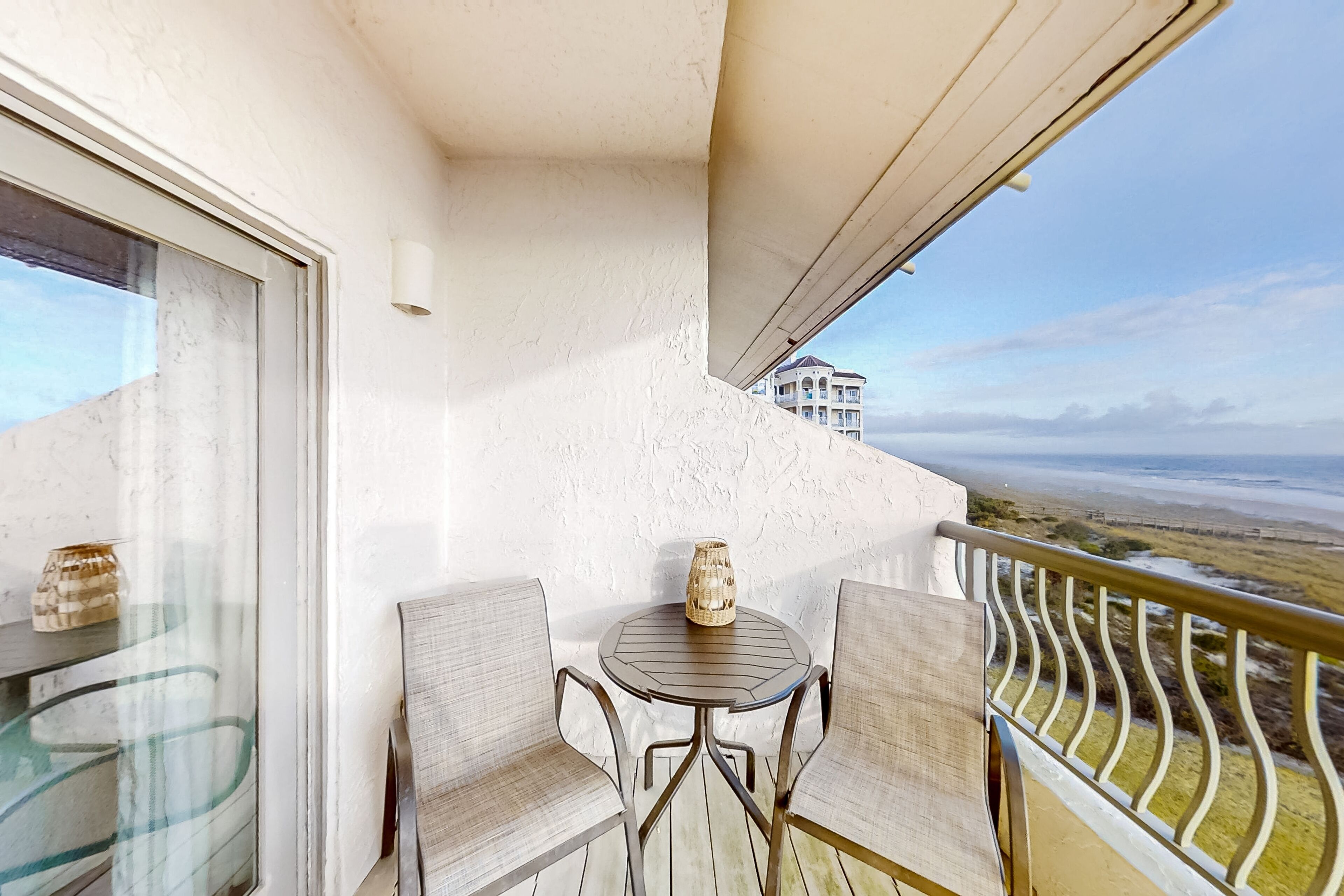 A balcony with two wicker chairs, a small table, and a decorative vase. A building is visible in the background.