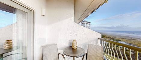 A balcony with two wicker chairs, a small table, and a decorative vase. A building is visible in the background.