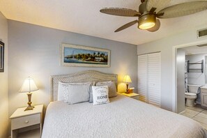 A bedroom with a bed, two bedside tables with lamps, a ceiling fan, a framed picture on the wall, and a bathroom visible through an open door. - 3rd-floor Condo W/fountain Views, Private Balcony & W/d, On-site Pool & hot Tubs (St. Augustine)