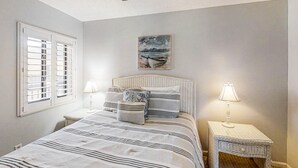 A bedroom with a bed, two nightstands, a ceiling fan, a window with shutters, and a painting of a boat on the wall. - 3rd-floor Condo W/fountain Views, Private Balcony & W/d, On-site Pool & hot Tubs (St. Augustine)