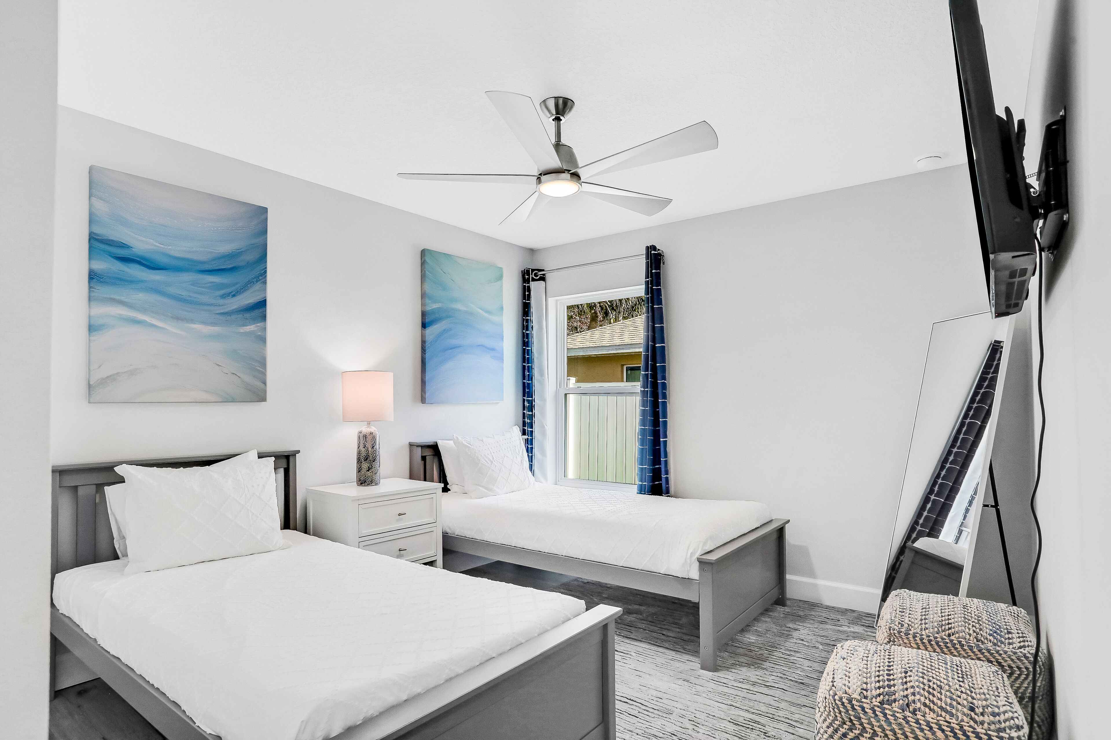 A modern bedroom with two beds, a ceiling fan, artwork, a nightstand, a lamp, and a TV mounted on the wall.