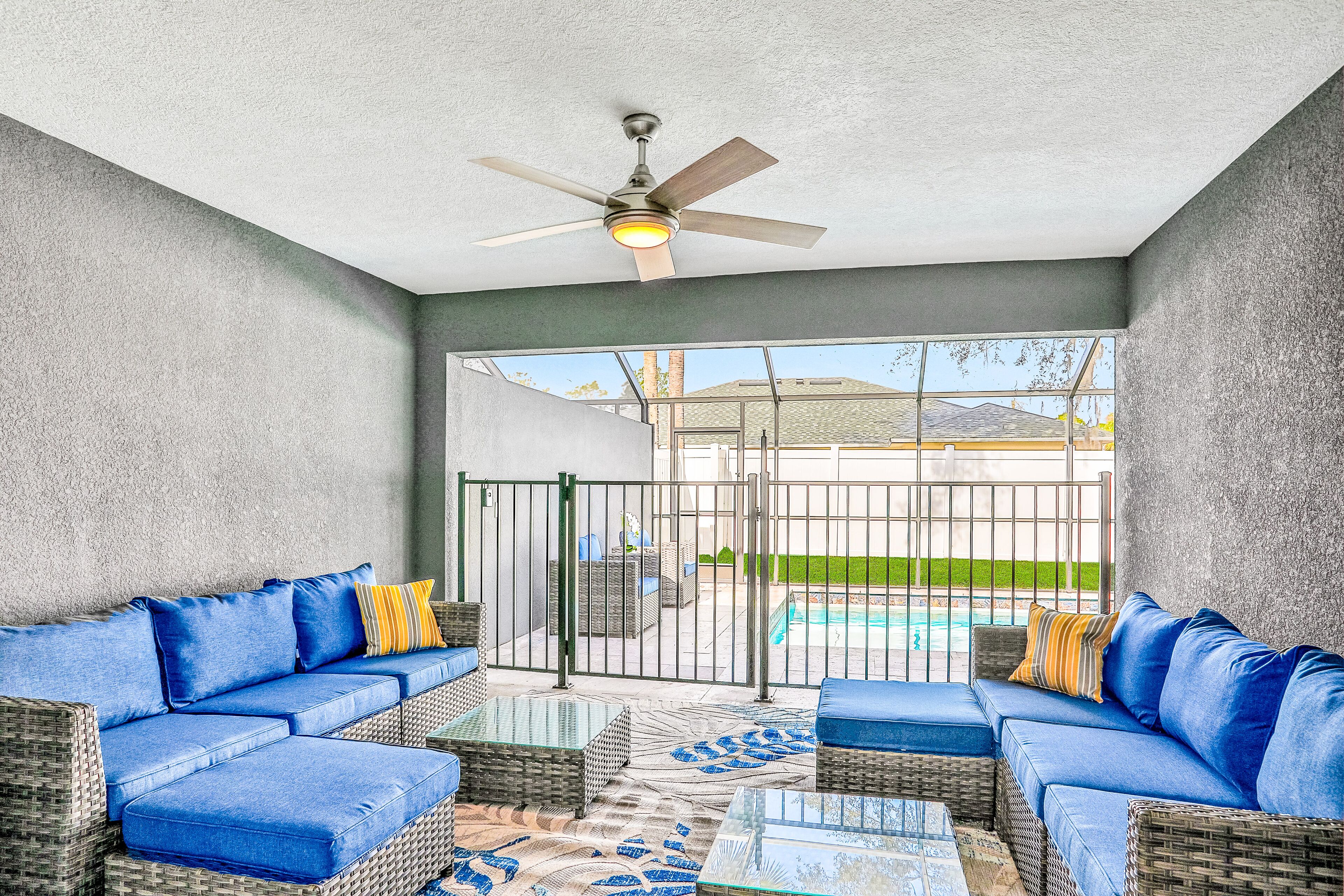 Un patio cubierto con muebles de mimbre, ventilador de techo y vista a un jardín con piscina.