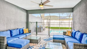 Un patio cubierto con muebles de mimbre, ventilador de techo y vista a un jardín con piscina.