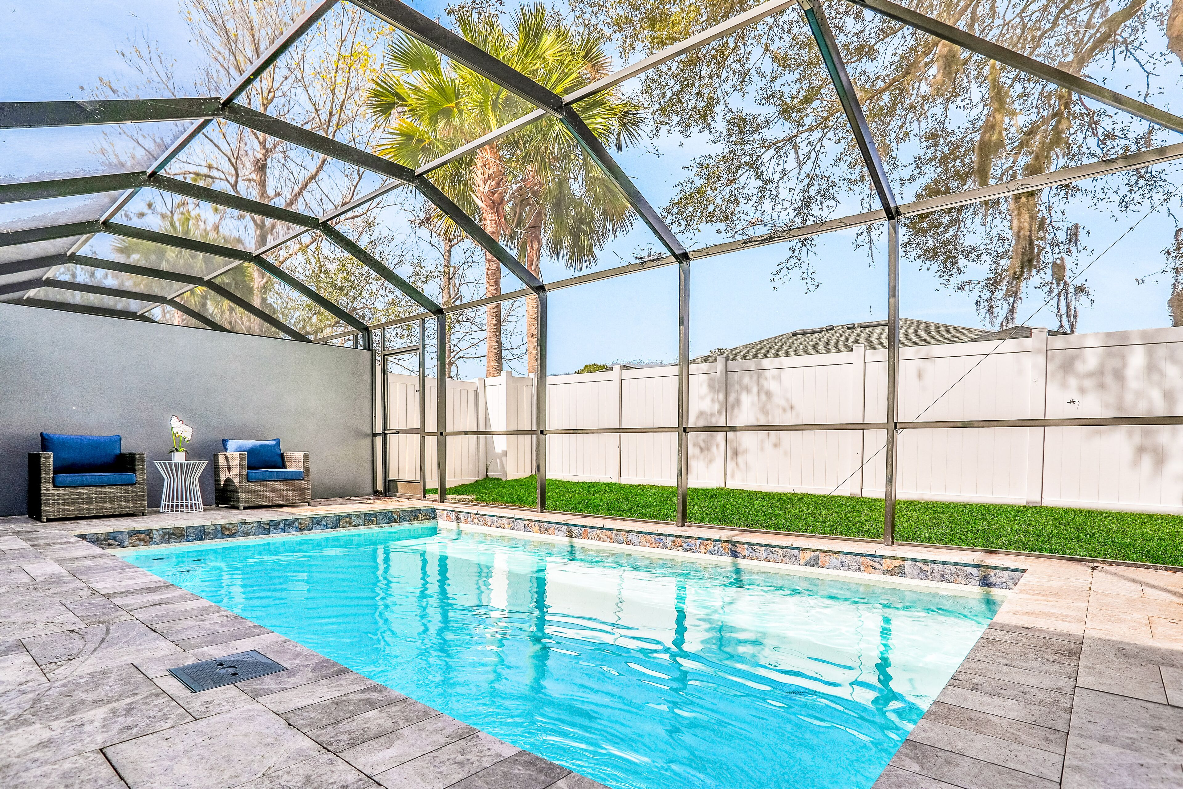 A swimming pool with a glass enclosure, surrounded by a patio area with outdoor furniture and a view of a grassy yard.