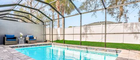 A swimming pool with a glass enclosure, surrounded by a patio area with outdoor furniture and a view of a grassy yard.