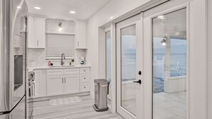 A modern kitchen with white cabinets, a stainless steel refrigerator, and a view of the sea through a glass door. - Beachfront Home w/ Private Lanai and Deck - Walk to the Beach (Flagler Beach)