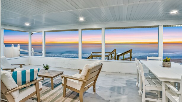 A covered outdoor seating area with a view of the ocean at sunset, featuring wooden furniture and a white table. - Beachfront Home w/ Private Lanai and Deck - Walk to the Beach (Flagler Beach)
