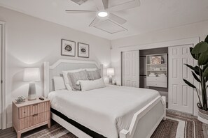 Een netjes ingerichte slaapkamer met een bed, bedlampen, een nachtkastje, een plafondventilator en een deur naar een badkamer.