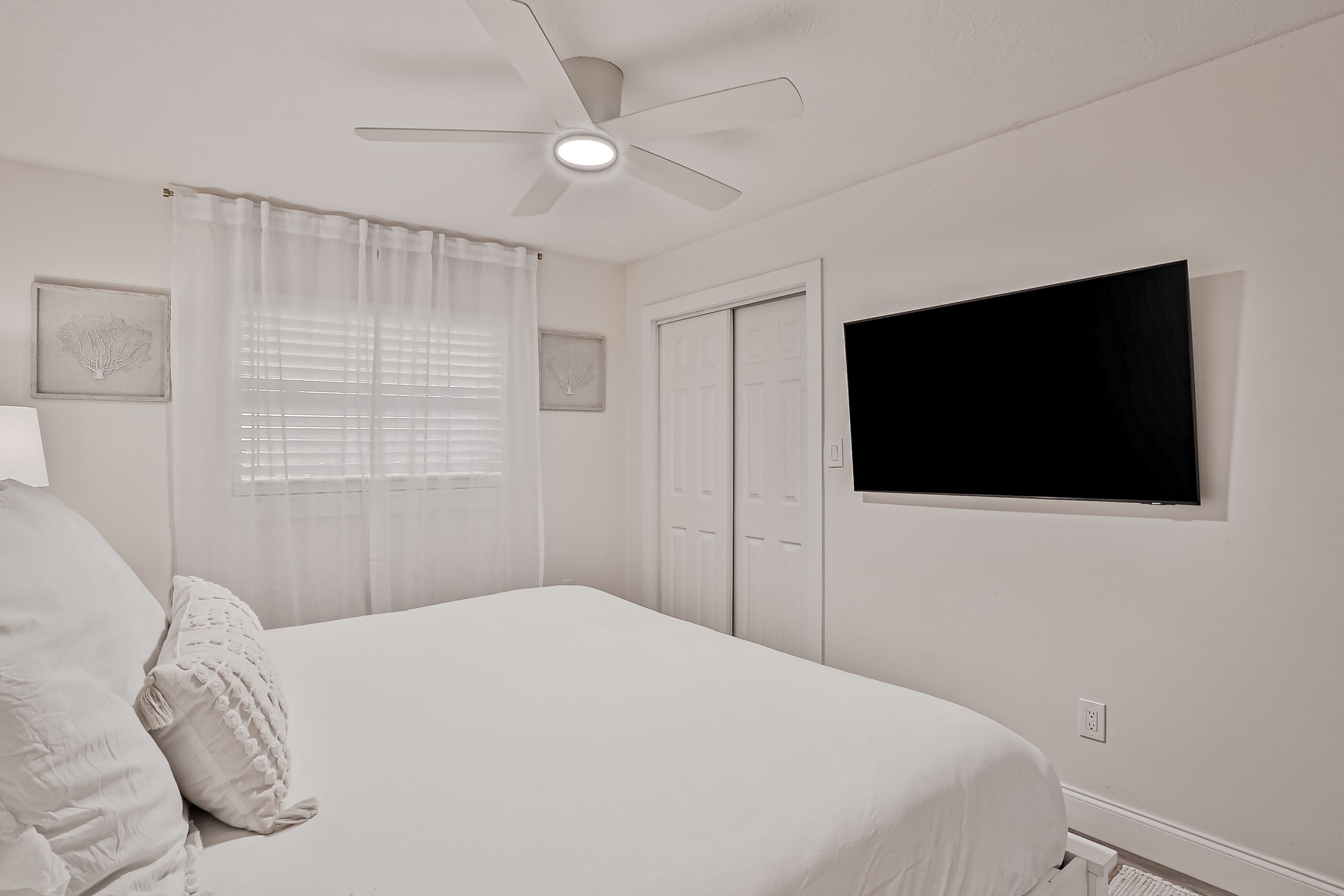 A bedroom with a bed, a ceiling fan, a TV mounted on the wall, and a window with sheer curtains.