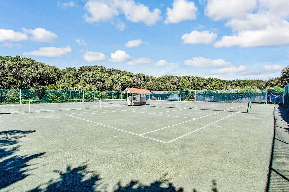 An outdoor tennis court with a net, surrounded by green fencing and trees in the background.