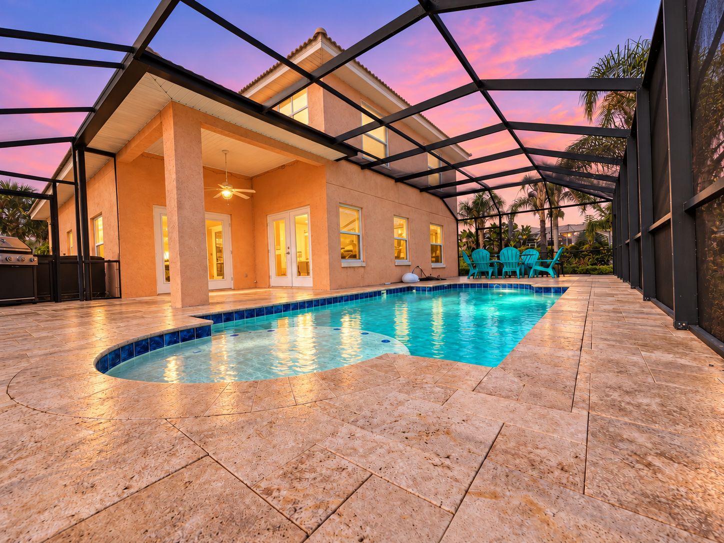 A covered swimming pool area with a house in the background, a patio with chairs, and a clear sky.