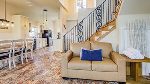 A spacious living area with a beige sofa, wooden dining table, and chairs. A staircase with a wrought iron railing leads to an upper level. - Coral Casa - Amazing Pool House Next to the Beach! 3 Min Walk! Awesome Pool! (Palm Coast)