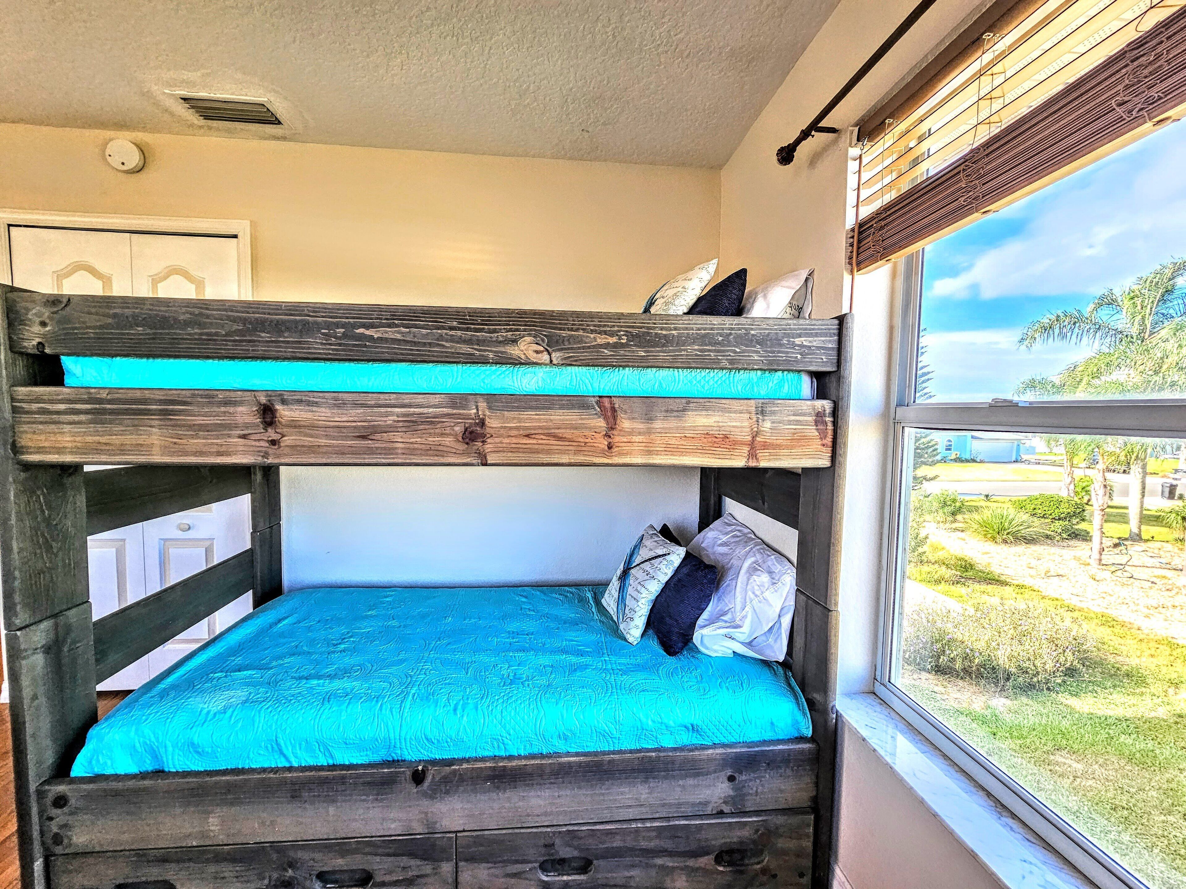Un lit superposé avec un matelas et des oreillers de couleur turquoise, une tête de lit en bois et une fenêtre donnant sur un paysage verdoyant et un bâtiment.