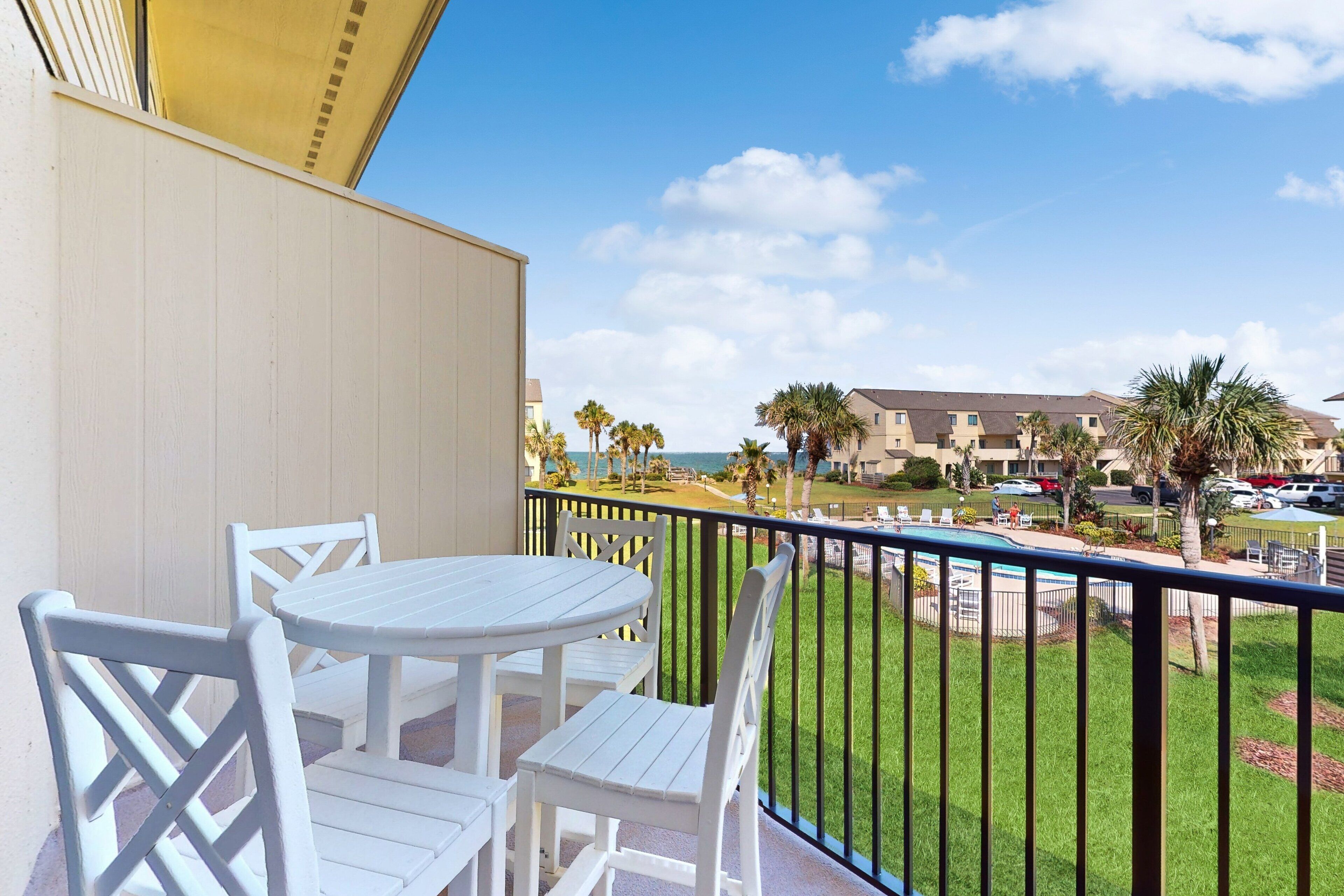 A balcony with white furniture, a view of a beach, and a building in the background.