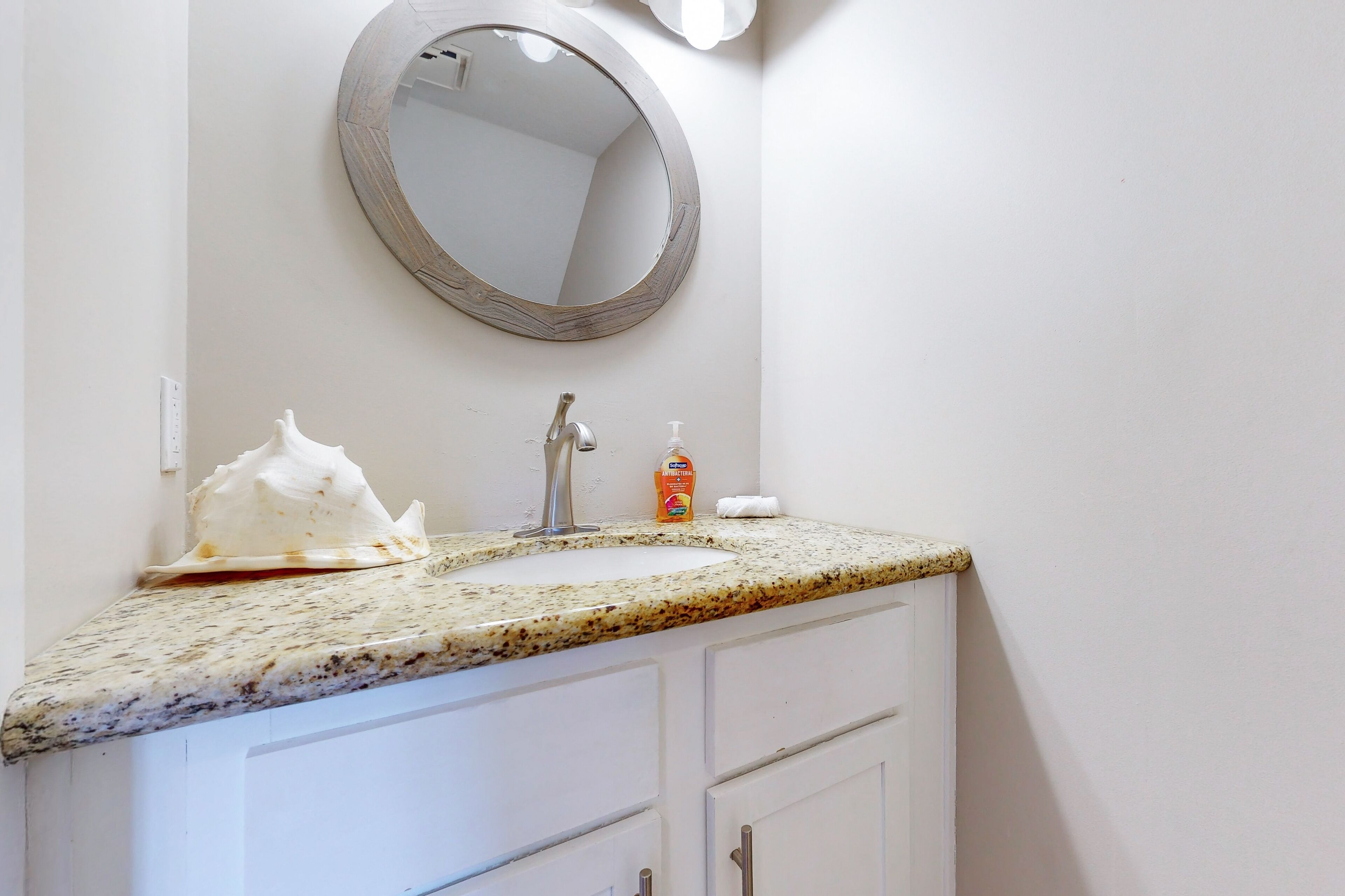Un baño con encimera de granito, un espejo redondo, un lavabo blanco y un dispensador de jabón.