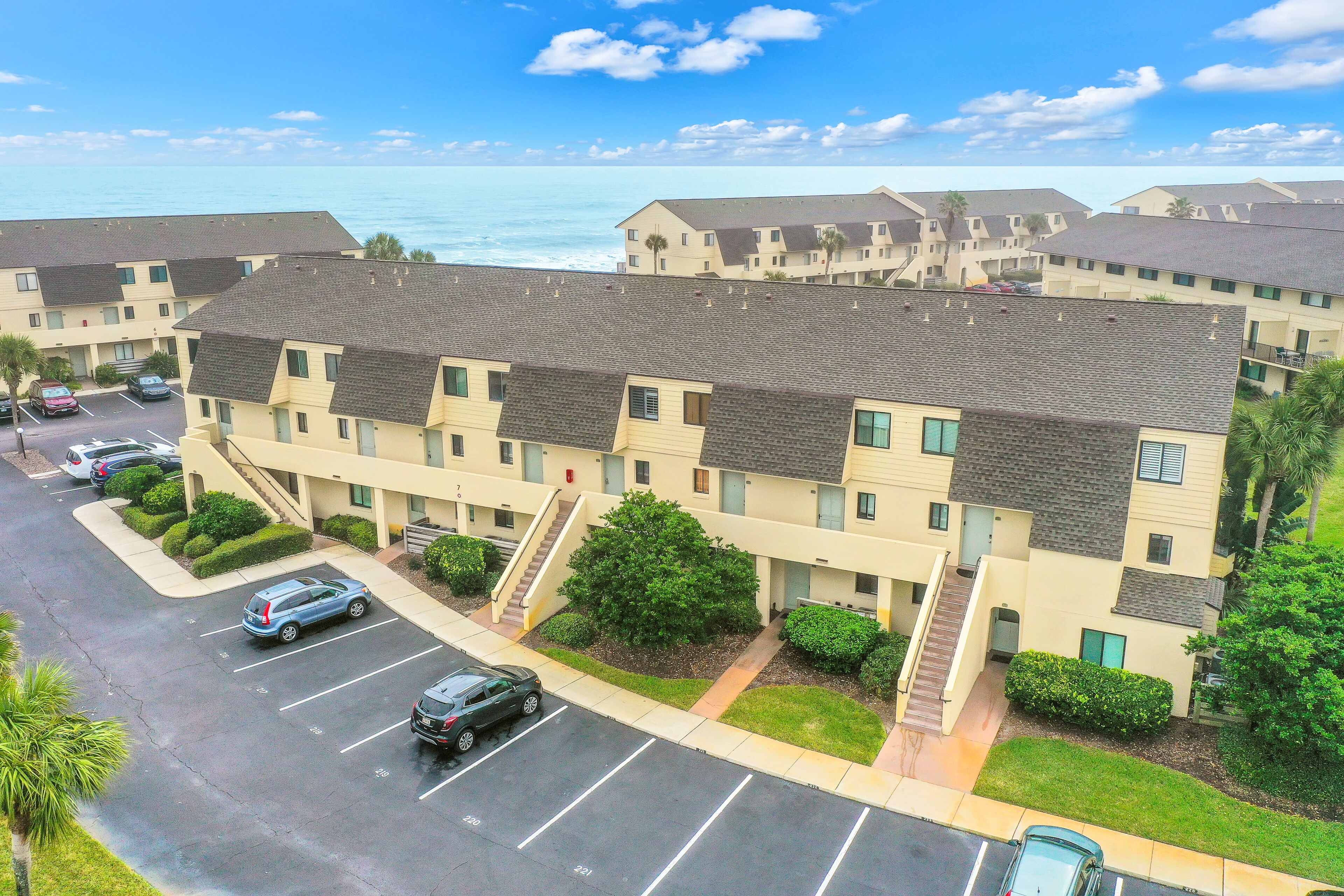A multi-story hotel with a parking lot, a staircase leading to the entrance, and a view of the ocean.