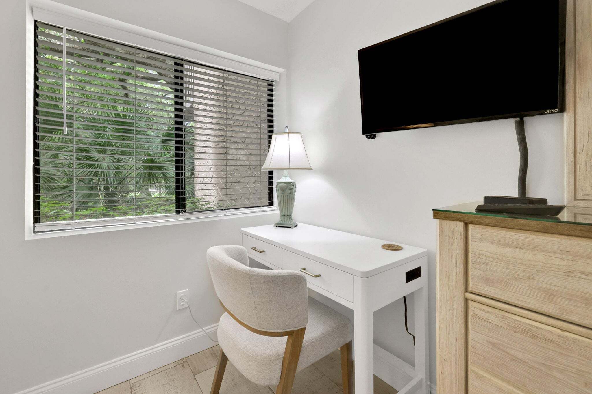 Una habitación con un escritorio blanco, una silla, una lámpara, un televisor y una ventana con persianas.