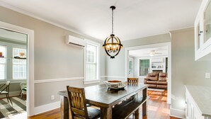 Uma área de refeições com mesa de madeira, cadeiras e um candelabro. Ao fundo, cozinha com armários brancos e um sofá de couro marrom.