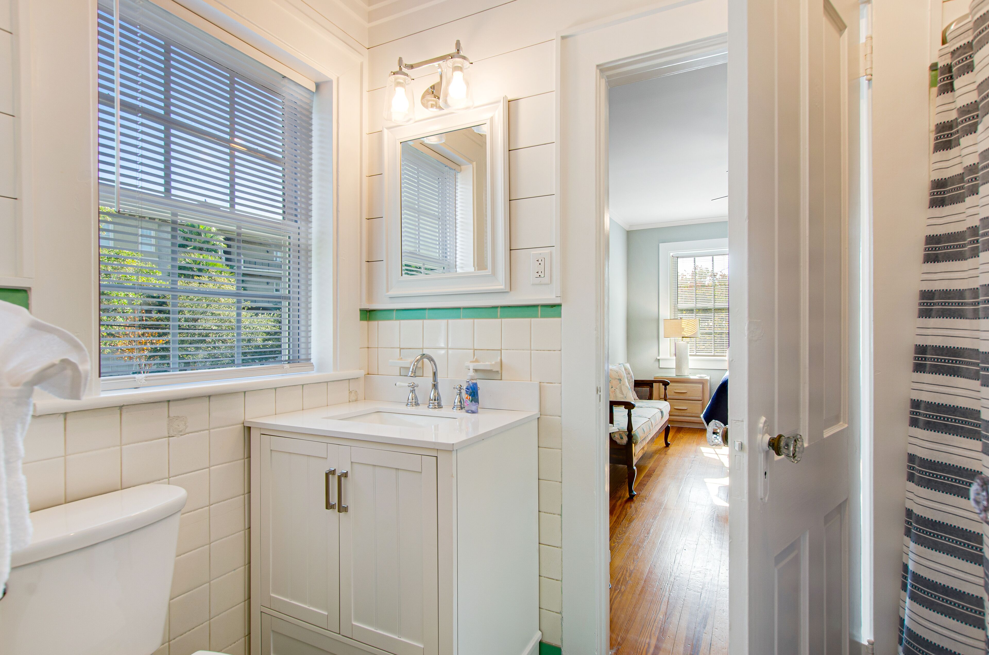 Une salle de bain équipée de toilettes, d’un lavabo et d’un miroir. Il y a une fenêtre avec des stores et une vue sur la verdure à l’extérieur.