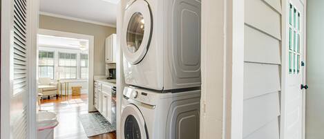 A laundry room with stacked washers and dryers, a wooden floor, and a view into a living area with a couch and a window.