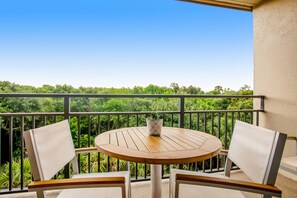 Une table ronde en bois, accompagnée de deux chaises blanches, est placée sur un balcon doté d’une rambarde en métal, d’où l’on peut admirer un paysage verdoyant et luxuriant.