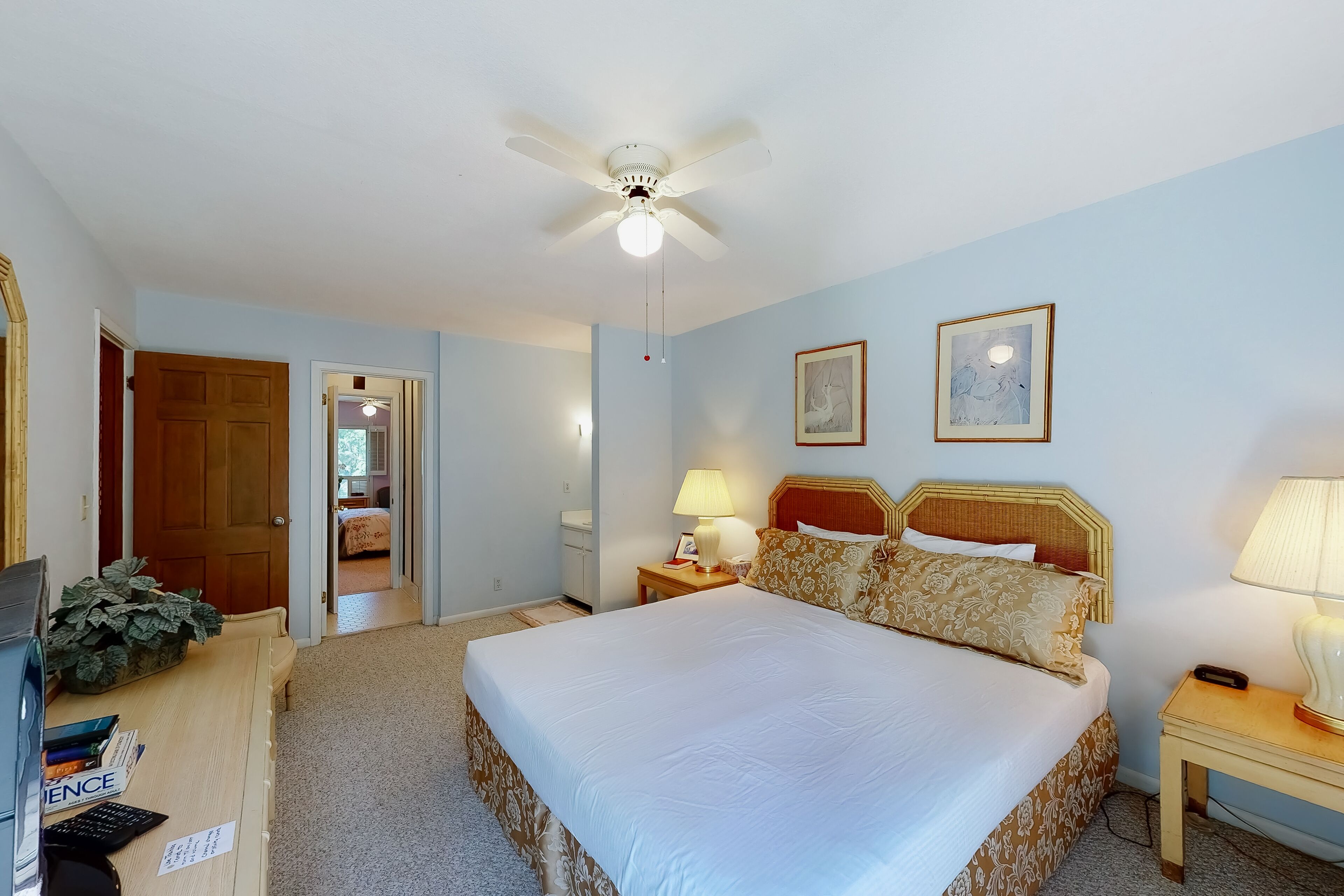 Une chambre d’hôtel comprenant un lit, un bureau, une chaise, un ventilateur de plafond, une table de chevet, une lampe, un tableau encadré et une porte donnant sur une autre pièce.