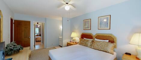 Une chambre d’hôtel comprenant un lit, un bureau, une chaise, un ventilateur de plafond, une table de chevet, une lampe, un tableau encadré et une porte donnant sur une autre pièce.