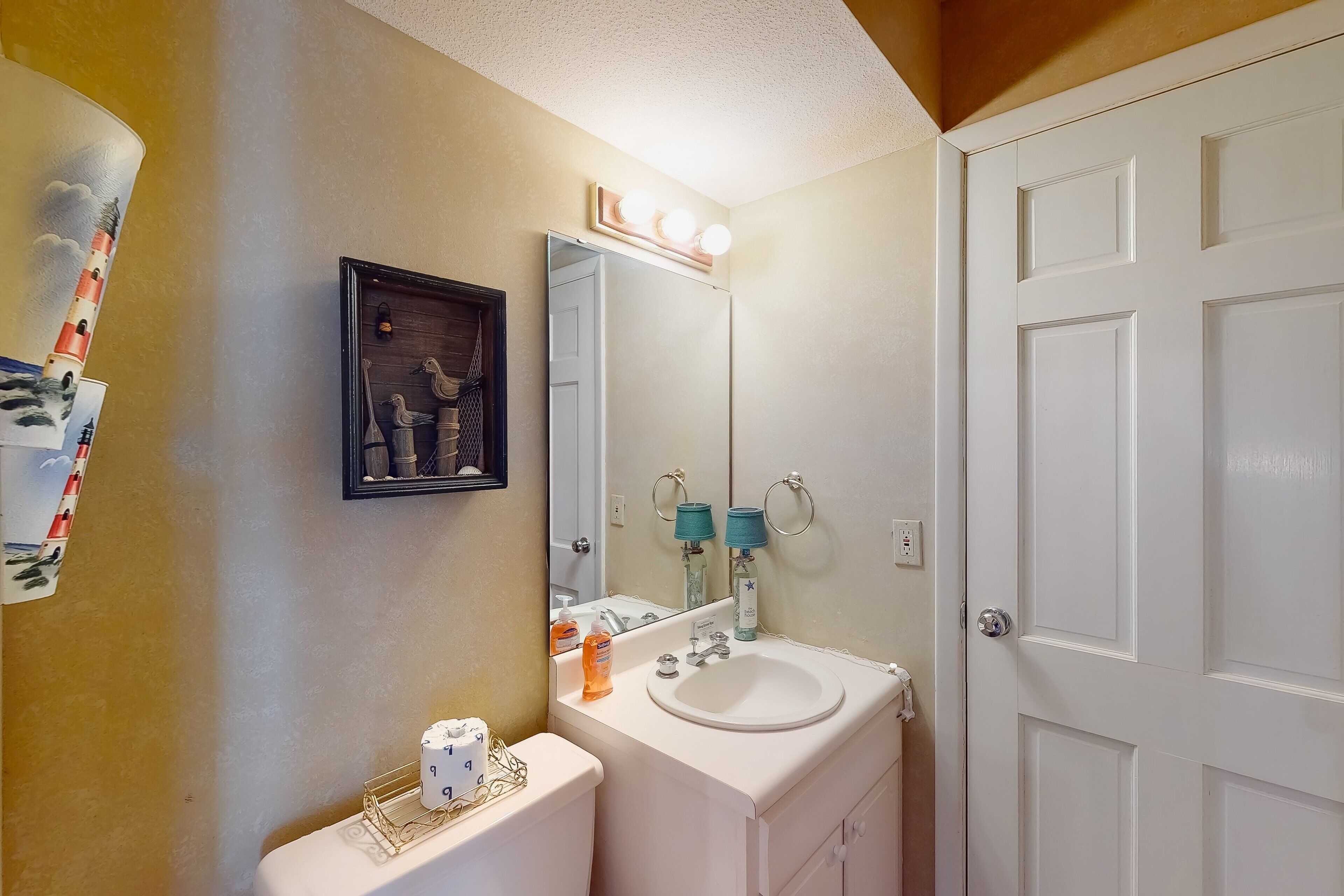 A bathroom with a white sink, a mirror with lights, a toilet, and a framed picture on the wall.