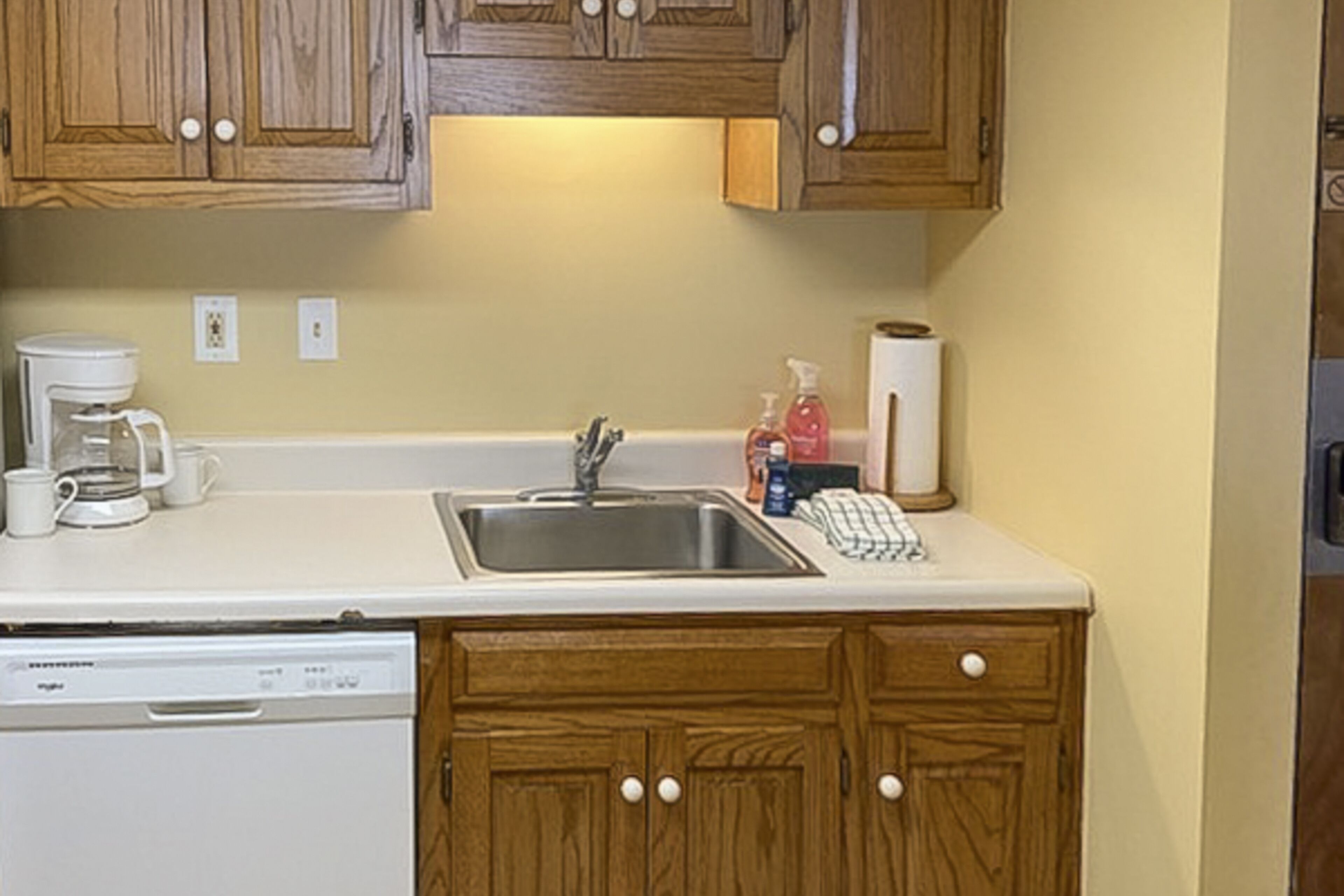 A kitchen with wooden cabinets, a white countertop, a coffee maker, a dishwasher, and a sink.