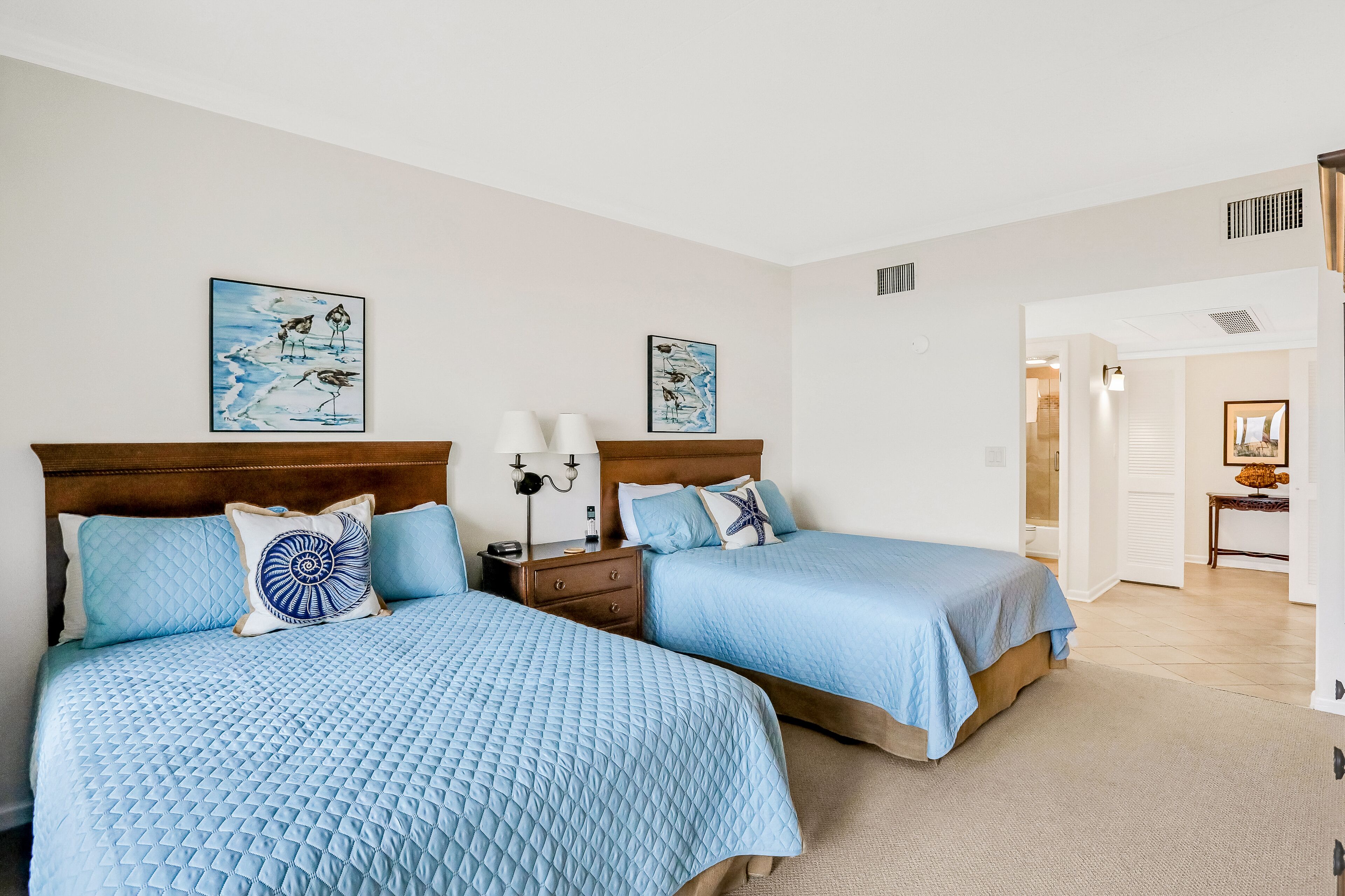 A bedroom with two beds, each with a blue quilted bedspread and matching pillows. There are two wall-mounted lamps on the nightstands.