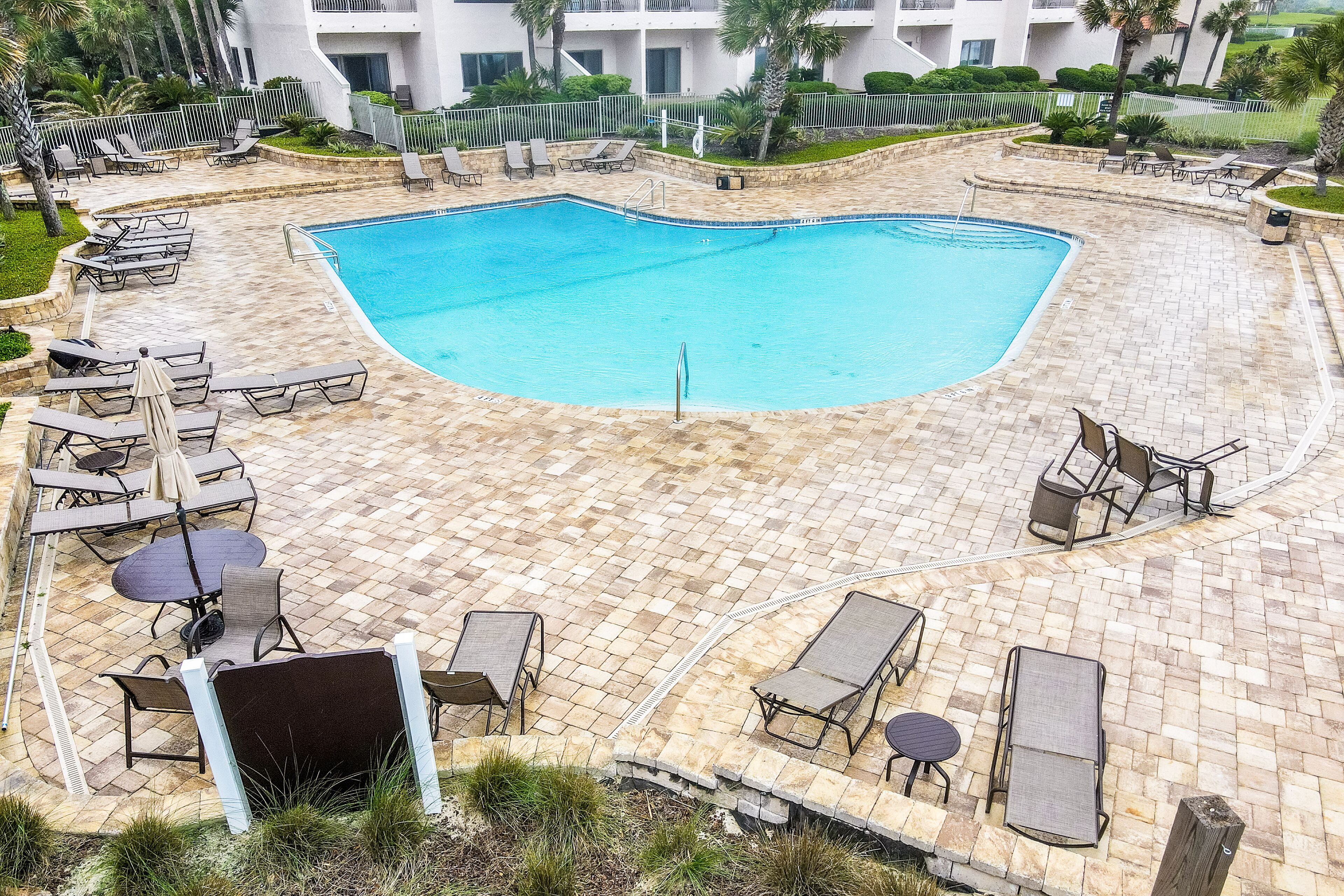 A swimming pool with lounge chairs and tables, surrounded by a brick patio and a fence.