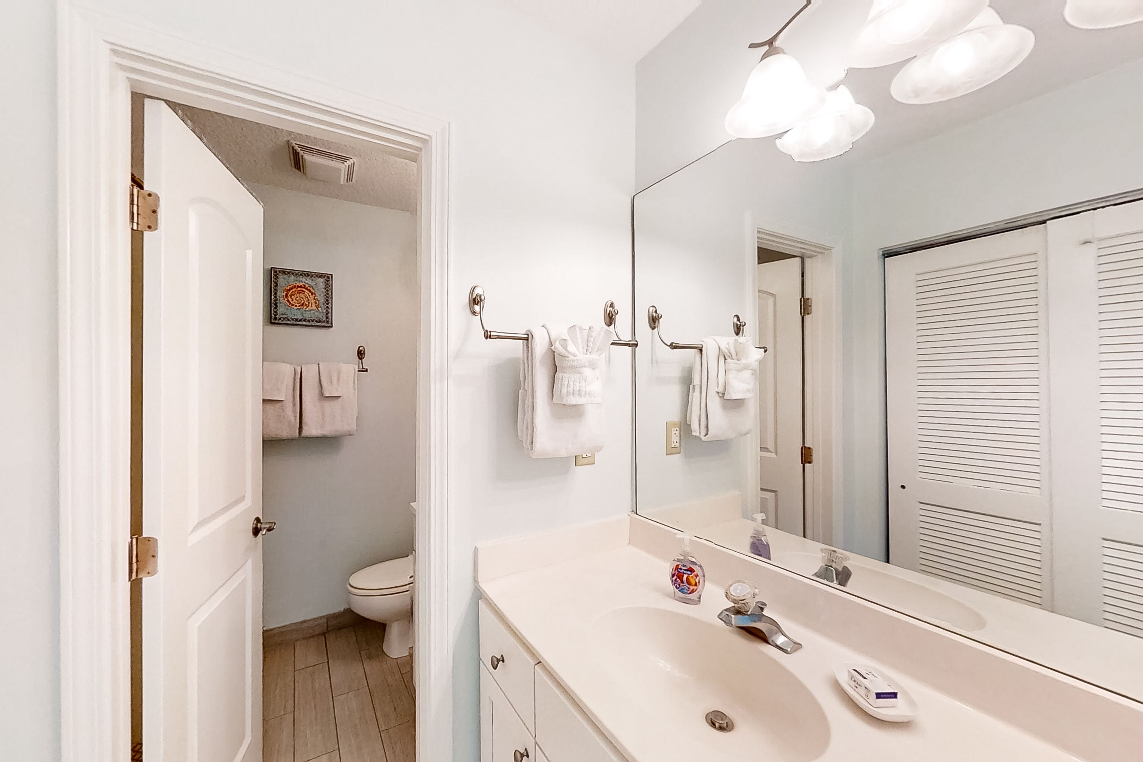 A bathroom with a white sink, a large mirror, and a toilet. There are towels hanging on a rack and a framed picture on the wall.