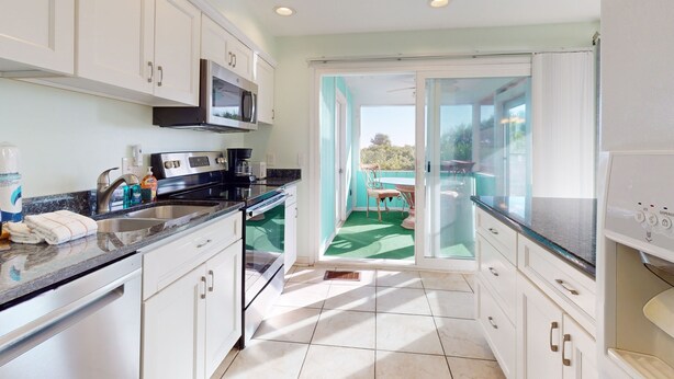 A modern kitchen with white cabinets, a stainless steel sink, and a microwave. There is a dining area with a table and chairs visible through a glass door leading to a balcony. - Ocean Paradise Bright Home With Spacious Deck, Beach Access, Central AC & Wifi (St. Augustine)