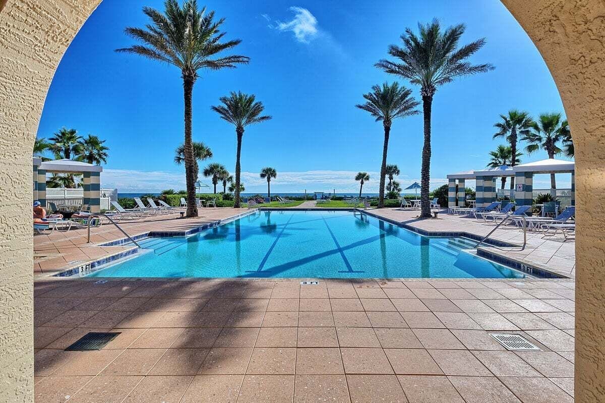 A swimming pool with clear blue water, surrounded by palm trees and lounge chairs, with a view of the ocean in the background.